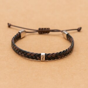 Peut inclure: Un bracelet tressé composé de brins alternés de cuir noir et marron foncé. Le bracelet comprend une perle centrale en métal argenté gravée d'un symbole infini contenant un petit cœur, ainsi que deux embouts de couleur argentée.