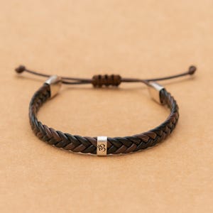 May include: A braided bracelet featuring alternating dark brown and black leather strands. The design includes a central silver-toned metal bead engraved with an Om symbol, two silver-toned end caps, and an adjustable sliding knot closure.