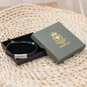 May include: A black braided cord bracelet featuring four silver-toned cylindrical beads engraved with the text 1985, a heart symbol, James, and Sara. The bracelet is presented in a black box with a dark green lid featuring a gold crown and diamond logo.