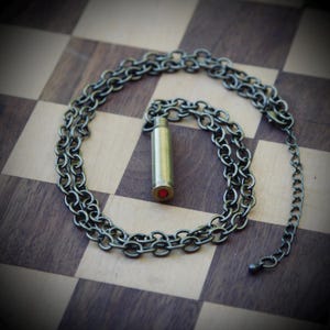 May include: A silver chain necklace with a bullet casing pendant. The bullet casing has a red jewel in the center.