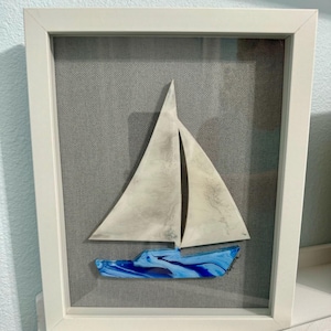 May include: A white framed picture with a blue and white sailboat cutout. The sailboat is on a gray fabric background.