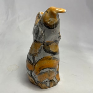 以下が含まれることがあります： 白、グレー、オレンジの抽象的な模様が施された猫のセラミック製置物。猫は座って、頭を後ろに傾け、耳を立てています。