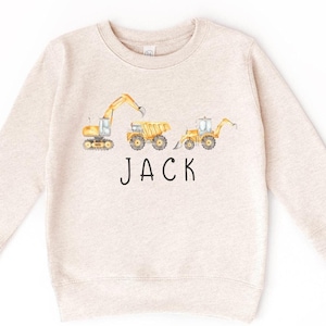 Peut inclure: Un sweat à manches longues beige avec un motif de véhicules de chantier et le nom "JACK" imprimé sur le devant.