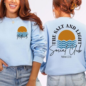 Könnte beinhalten: Hellblaues Sweatshirt mit einem Sonnen- und Wellen-Motiv und dem Text "Matthew 5". Auf der Rückseite des Sweatshirts steht "The Salt and Light Social Club" mit einem Sonnen- und Wellen-Design.