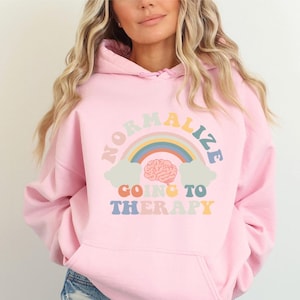 May include: Pink hoodie with a rainbow and brain graphic that says "Normalize Going to Therapy".