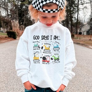 Peut inclure: Sweat-shirt blanc avec un graphique de véhicules de chantier et le texte "GOD SAYS I AM..." avec des versets bibliques listés sous chaque véhicule.