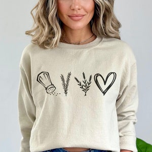 May include: A beige sweatshirt with a black graphic of a chef's hat, herbs, and a heart. The graphic says "YUM".