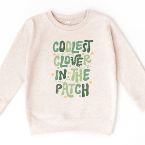May include: A beige sweatshirt with the text "COOLEST CLOVER IN THE PATCH" in green and yellow lettering. The sweatshirt has long sleeves and a crew neck. The design is centered on the front of the garment.