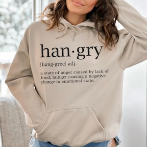 Könnte beinhalten: Ein beiger Hoodie mit dem Wort "han-gry" definiert als "ein Zustand der Wut, der durch einen Mangel an Nahrung verursacht wird; Hunger, der eine negative Veränderung des emotionalen Zustands verursacht."