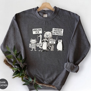 May include: Dark grey sweatshirt featuring cartoon animals holding signs with text including "Abolish ICE" and "Defund ICE". The sweatshirt is displayed on a wooden hanger.