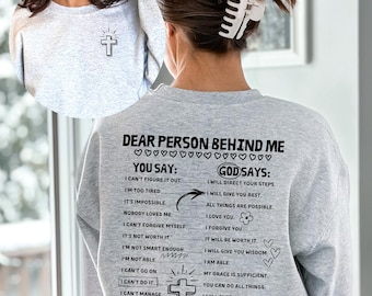 May include: Grey sweatshirt with a cross graphic and text that reads "Dear person behind me. You say: I can't figure it out. I'm too tired. It's impossible. Nobody loves me. I can't forgive myself. It's not worth it. I'm not smart enough. I'm not able. I can't go on. I can't do it. I can't manage. I'm afraid. I feel all alone. God says: I will direct your steps. I will give you rest. All things are possible. I love you. I forgive you. It will be worth it. I will give you wisdom. I am able. My grace is sufficient. You can do all things. I will supply all you need. I have not given you fear. I will never leave you."