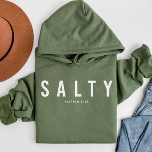 Może przedstawiać: Zielona bluza z kapturem z nadrukiem "SALTY" w kolorze białym na przodzie. Pod napisem "SALTY" znajduje się tekst "MATTHEW 5:13".