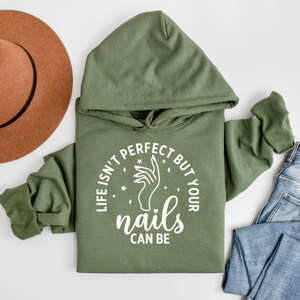 Op de afbeelding: Een groen hoodie met de tekst "Life isn't perfect but your nails can be" in witte letters. De tekst staat in een cirkel met een hand die een vijl vasthoudt in de cirkel.