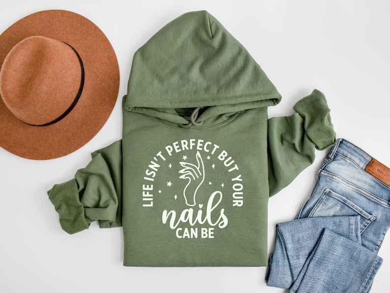 Op de afbeelding: Een groen hoodie met de tekst "Life isn't perfect but your nails can be" in witte letters. De tekst staat in een cirkel met een hand die een vijl vasthoudt in de cirkel.
