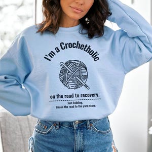 K&ouml;nnte beinhalten: Hellblaues Sweatshirt mit dem Text "I'm a Crochetholic on the road to recovery. Just kidding. I'm on the road to the yarn store." Die Grafik zeigt einen Wollkn&auml;uel und H&auml;kelnadeln.