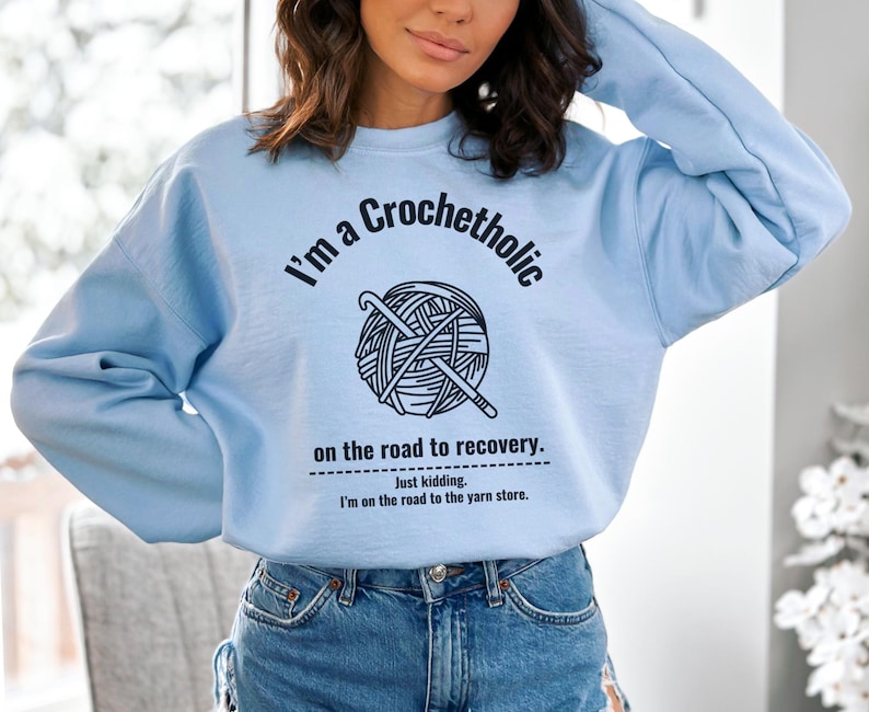 K&ouml;nnte beinhalten: Hellblaues Sweatshirt mit dem Text "I'm a Crochetholic on the road to recovery. Just kidding. I'm on the road to the yarn store." Die Grafik zeigt einen Wollkn&auml;uel und H&auml;kelnadeln.