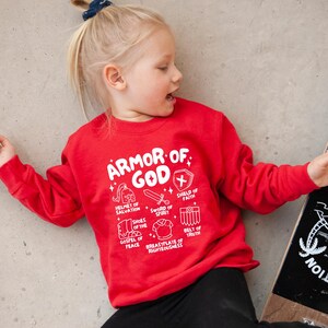 May include: Red sweatshirt with a white graphic of the Armour of God. The graphic includes a helmet, sword, shield, shoes, breastplate, and belt. The text reads "Armour of God" and lists the different pieces of armour.