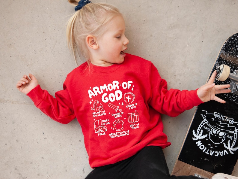 May include: Red sweatshirt with a white graphic of the Armour of God. The graphic includes a helmet, sword, shield, shoes, breastplate, and belt. The text reads "Armour of God" and lists the different pieces of armour.