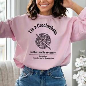 K&ouml;nnte beinhalten: Hellrosa Sweatshirt mit dem Text "I'm a Crochetholic" &uuml;ber einer Grafik aus Garn und H&auml;kelnadeln. Darunter steht der Text: "on the road to recovery. Just kidding. I'm on the road to the yarn store."