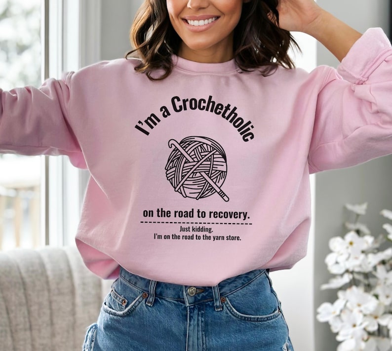 K&ouml;nnte beinhalten: Hellrosa Sweatshirt mit dem Text "I'm a Crochetholic" &uuml;ber einer Grafik aus Garn und H&auml;kelnadeln. Darunter steht der Text: "on the road to recovery. Just kidding. I'm on the road to the yarn store."