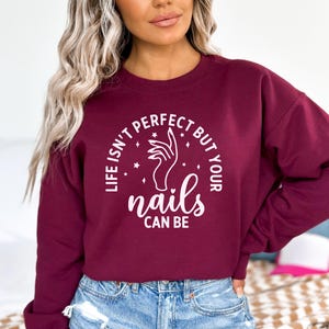 Op de afbeelding: Bordeauxkleurige trui met een witte afbeelding met de tekst "Het leven is niet perfect, maar je nagels kunnen dat wel zijn". De afbeelding bevat ook een hand met gemanicuurde nagels en sterren.