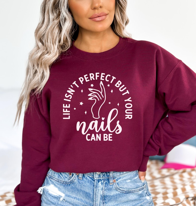 Op de afbeelding: Bordeauxkleurige trui met een witte afbeelding met de tekst "Het leven is niet perfect, maar je nagels kunnen dat wel zijn". De afbeelding bevat ook een hand met gemanicuurde nagels en sterren.