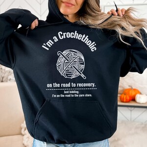 K&ouml;nnte beinhalten: Schwarzer Kapuzenpullover mit dem Text "I'm a Crochetholic" &uuml;ber einer Garnkugel und H&auml;kelhaken-Grafik. Unter der Grafik steht der Text "on the road to recovery. Just kidding. I'm on the road to the yarn store."