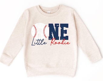 May include: A beige long-sleeved sweatshirt with a baseball graphic and the text "One Little Rookie" in blue and red.