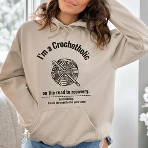 May include: A beige hoodie with a front pocket and drawstring hood. The front features the text "I'm a Crochetholic" above a graphic of yarn and crochet hooks, with the text "on the road to recovery." and "Just kidding. I'm on the road to the yarn store."
