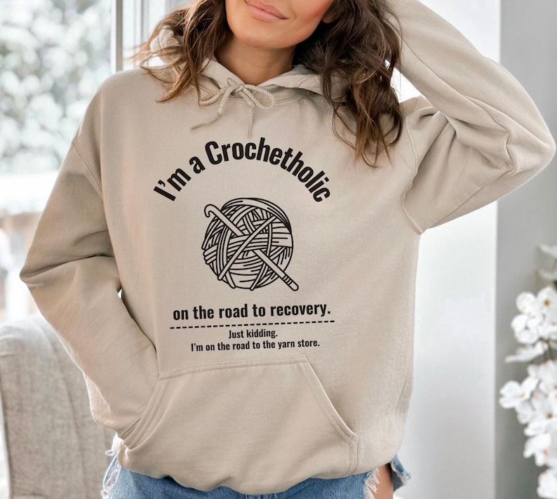 K&ouml;nnte beinhalten: Ein beigefarbener Kapuzenpullover mit einer Vordertasche und einem Kordelzug. Auf der Vorderseite steht der Text "I'm a Crochetholic" &uuml;ber einer Grafik von Garn und H&auml;kelnadeln, mit dem Text "on the road to recovery." und "Just kidding. I'm on the road to the yarn store."