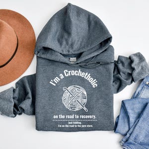 K&ouml;nnte beinhalten: Ein grauer Kapuzenpullover mit dem Text "I'm a Crochetholic" und einer Garnkn&auml;uel-Grafik. Der Text "on the road to recovery" und "Just kidding. I'm on the road to the yarn store" sind ebenfalls auf der Vorderseite aufgedruckt. Ein brauner Hut und blaue Jeans sind ebenfalls im Bild.