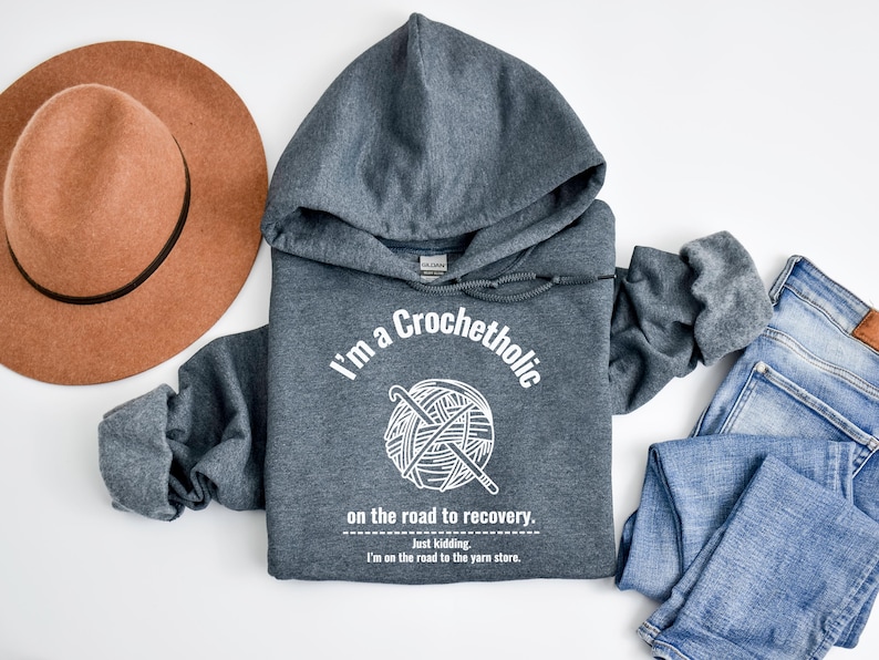 K&ouml;nnte beinhalten: Ein grauer Kapuzenpullover mit dem Text "I'm a Crochetholic" und einer Garnkn&auml;uel-Grafik. Der Text "on the road to recovery" und "Just kidding. I'm on the road to the yarn store" sind ebenfalls auf der Vorderseite aufgedruckt. Ein brauner Hut und blaue Jeans sind ebenfalls im Bild.