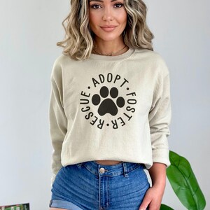 May include: A beige sweatshirt with a black paw print graphic and the text "Adopt. Foster. Rescue." printed in a circle around the paw print.