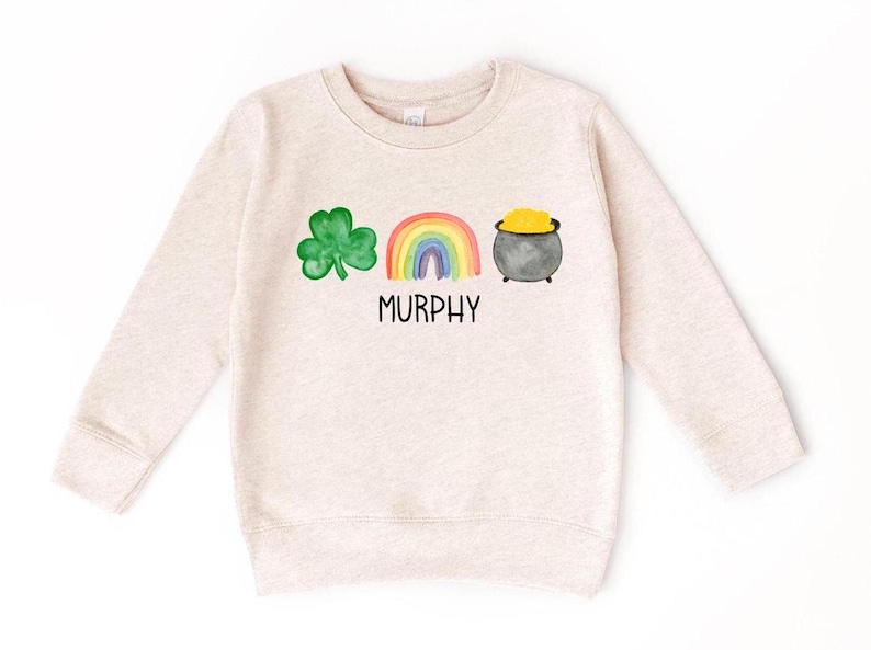 May include: A light beige sweatshirt with a green shamrock, a rainbow, and a pot of gold. The text "MURPHY" is printed below the design.