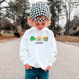 May include: A white sweatshirt with a green shamrock, rainbow, and pot of gold graphic. The text "JHONSON" is printed below the graphic. A child is wearing the sweatshirt and a black and white checkered hat.