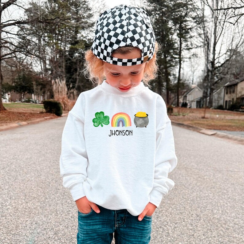 May include: A white sweatshirt with a green shamrock, rainbow, and pot of gold graphic. The text "JHONSON" is printed below the graphic. A child is wearing the sweatshirt and a black and white checkered hat.