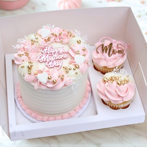 May include: A white cake with pink frosting and gold sprinkles, decorated with a "Happy Mother's Day" message. The cake is in a white box with two pink cupcakes decorated with gold sprinkles and "Mom" toppers.