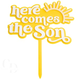 Pode incluir: Um topo de bolo amarelo com a frase "here comes the Son" em branco, com um desenho de sol. O topo tem uma haste longa e fina para inserir em um bolo ou outra sobremesa.