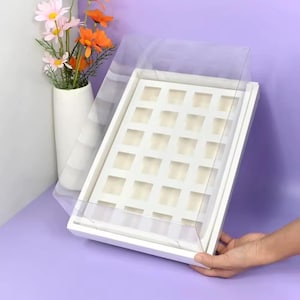 May include: White cardboard box with a clear plastic lid. The box has 36 square compartments for holding small items.