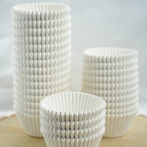 May include: A collection of white, pleated paper cupcake liners. The liners are stacked in varying heights, with the tallest stack on the left and a smaller stack on the right. A few liners are stacked in the foreground.