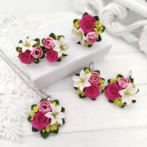 Puede incluir: Un conjunto de joyas con diseños florales. El conjunto incluye un collar, pendientes y un anillo, todos adornados con rosas rosas, lirios blancos y hojas verdes. Las joyas están sobre un fondo blanco.