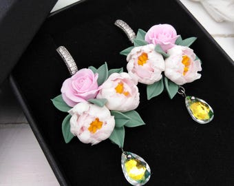 May include: A pair of floral earrings featuring pink and white peony and rose designs with green leaves. Each earring has a clear, teardrop-shaped crystal pendant and a silver-colored, curved ear wire.