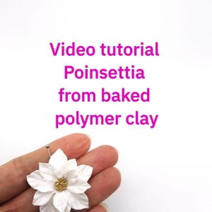 May include: A handmade white poinsettia flower with a gold center, held by a hand. The flower is made from baked polymer clay and is the subject of a video tutorial. Text in the image reads: "Video tutorial Poinsettia from baked polymer clay."
