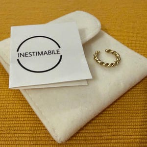 May include: A gold-coloured ear cuff with a twisted rope design sits on a white pouch. A white card with the word "INESTIMABILE" in a black semi-circle is also present. The background is a textured yellow surface.