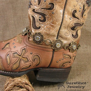 May include: Brown leather cowboy boot with intricate floral designs and silver studs. A gold chain with decorative bullet charms wraps around the boot.