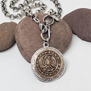 May include: Silver-toned necklace with a large, round pendant. The pendant features a gold-colored center with an eagle design and the inscription "Astra Devs Spes". The necklace has a lobster clasp and a chain with large, round links.