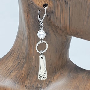 May include: A single silver earring with a lever back closure. The earring features a white pearl bead, a silver ring, and a decorative spoon-shaped charm. The charm has an ornate floral design at the bottom. The earring is shown against a wooden background.
