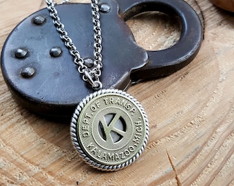 May include: A silver chain necklace with a round pendant. The pendant is a brass replica of a transit token with the words "DEPT OF TRANS" and "KANSAS CITY, MO" around the edge. The letter "K" is in the center of the token.