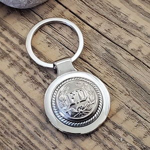 May include: Silver key chain with a round metal charm featuring the letters "FD" surrounded by a wreath design.