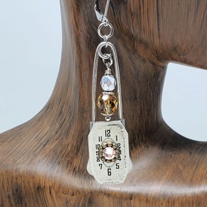 Puede incluir: Pendientes de plata con un dije de esfera de reloj vintage, un strass transparente y cuentas marrones y plateadas. La esfera del reloj dice "SWITZERLAND" en la parte inferior.